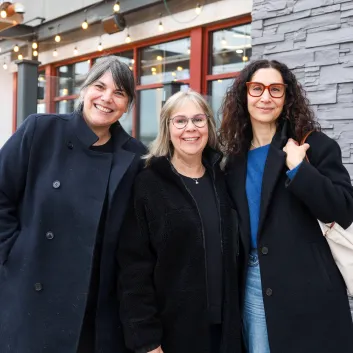 Three women smiling together outdoors, wearing dark coats on a cloudy day.