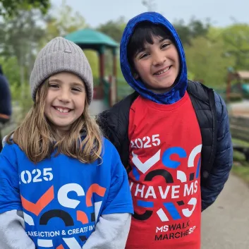 Two smiling children in numbered race bibs at an outdoor charity walk event.