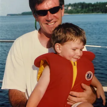Larry in sunglasses holding his son Glen in a red life jacket on a boat.