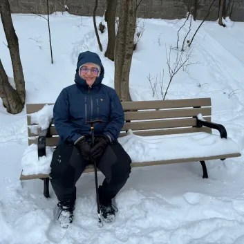 Ksenia in winter gear sitting on a snow-covered park bench outside.