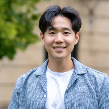 Dr. Paul Yoo is wearing a white t-shirt with a light blue blazer. He's smiling and standing in front of a building and trees.