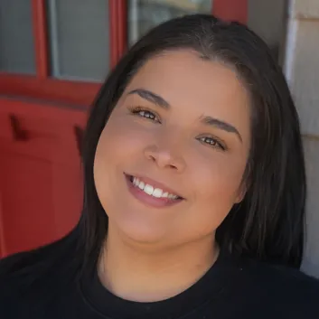 Rachel smiling in a black "quietly powerful" sweatshirt, standing near a red door.