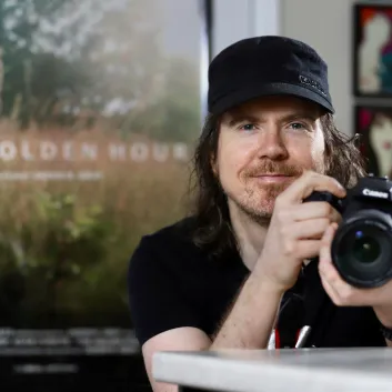 Jason wearing a black t-shirt and hat, holding a camera with a poster that reads "The Golden Hour" behind him.