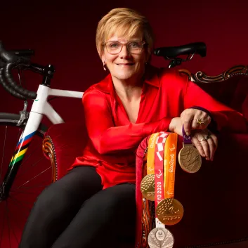 Carol in a red top and black pants sitting in a red armchair, holding medals, with a road bike behind her. 