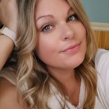  Michelle with long wavy blonde hair resting her head on her raised arm, wearing a white top.